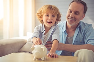 An older man is holding his grandson while the young boy puts money into a piggy bank on the table