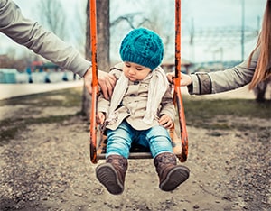 A toddler is on a swing with an arm from each parent on either side of the swing