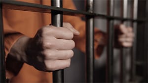 A man wearing an orange prison jumpsuit who has been charged with a felony grips the bars of his jail cell