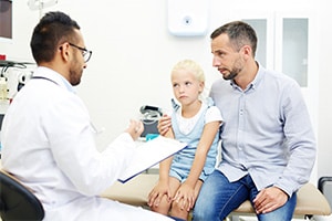 A father sitting next to his young daughter in a doctor's exam room while a doctor speaks to them