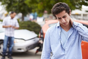 A young man rubs his neck in pain while standing in front of two cars that have crashed into each other