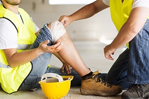 A man wearing construction gear bends down to help another man in construction gear who has injured his knee